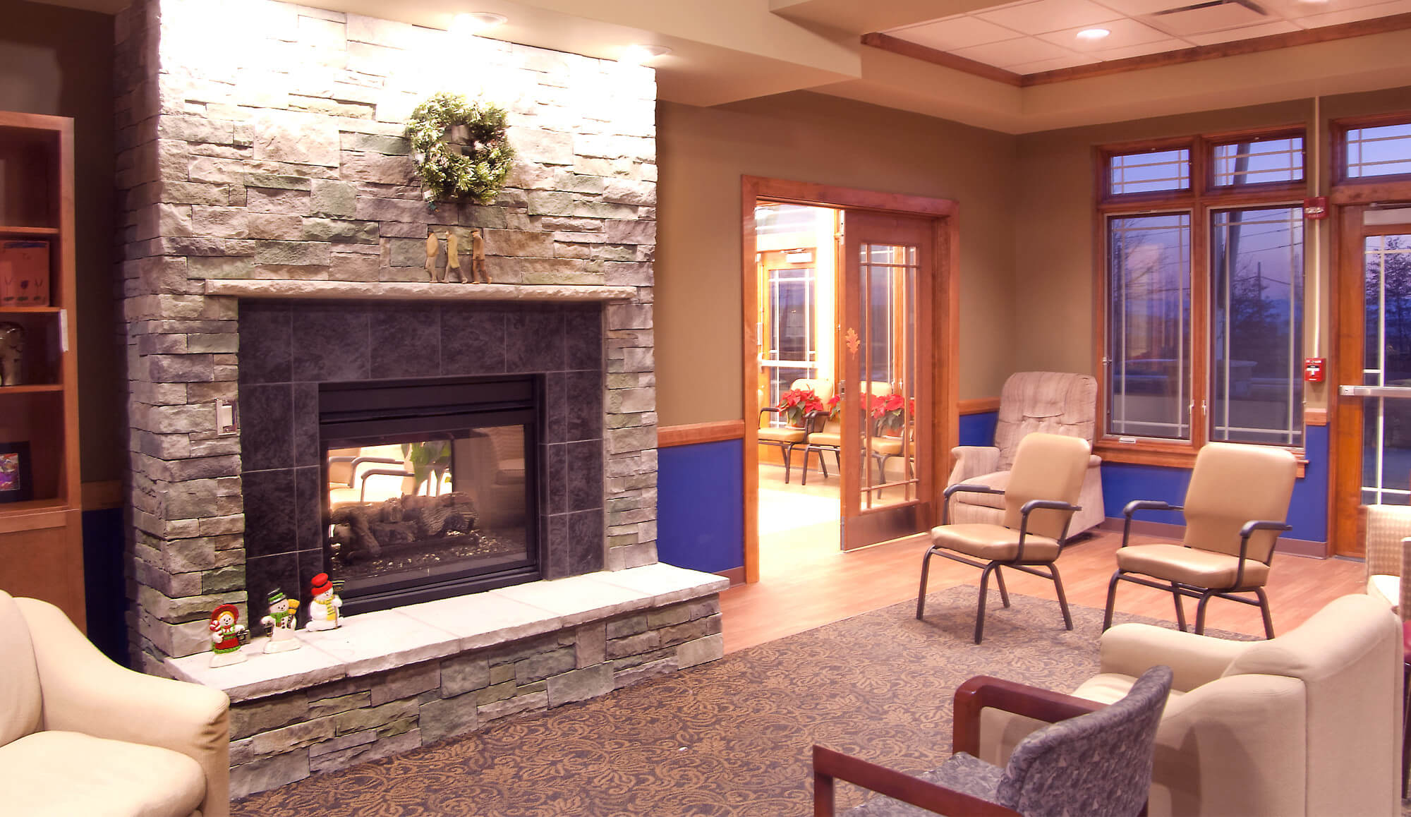 A room with multiple chairs, a fireplace with a wreath above it, and many windows.