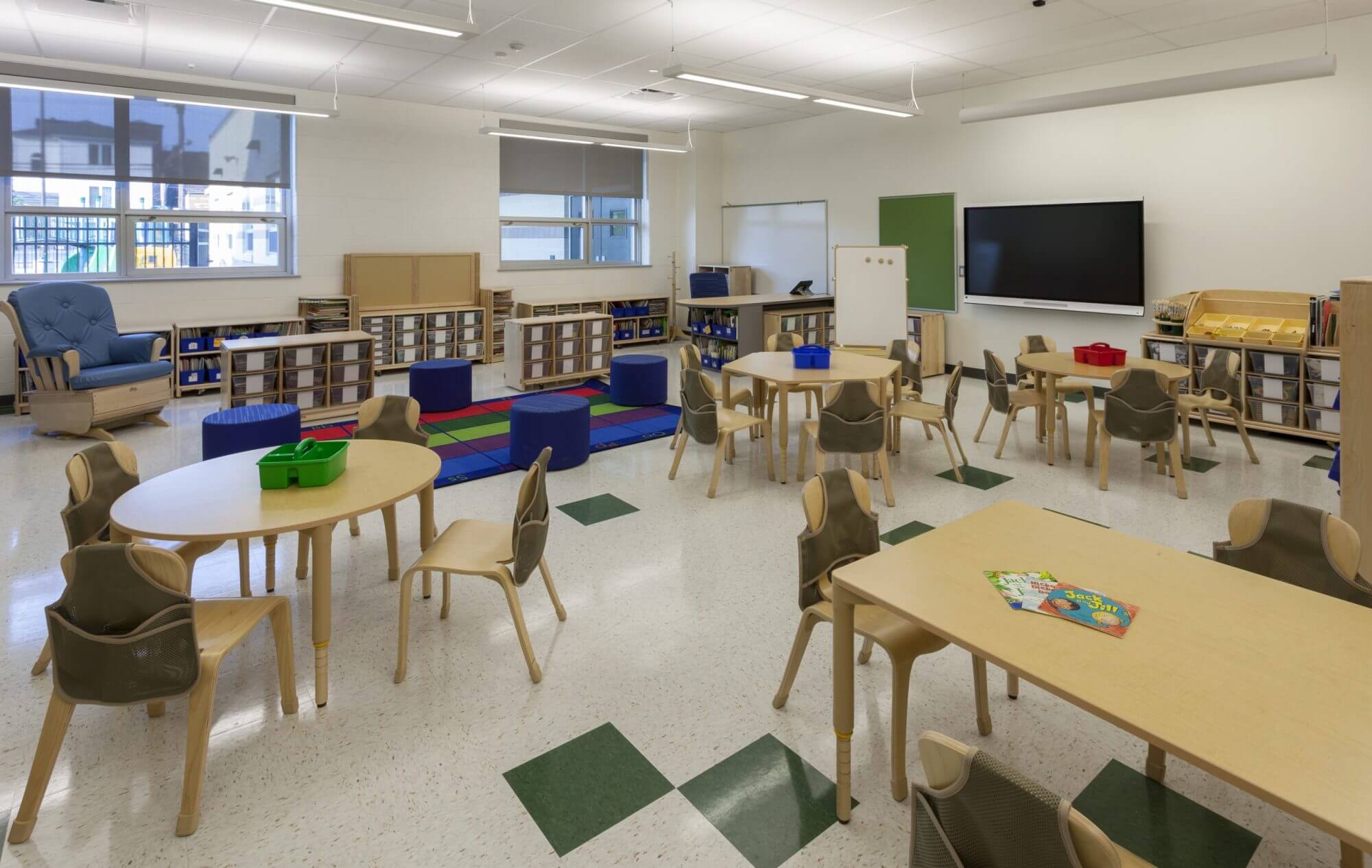 Inside a classroom with multiple tan desks, various shaped chairs, a book area, and a large screen mounted on the wall.