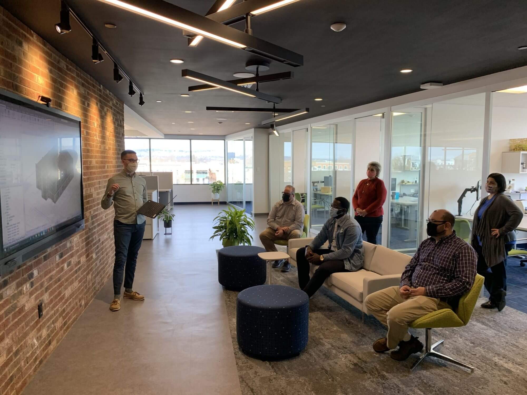 Five people wearing facemasks listening to another person wearing a face mask give a presentation at an office with a brick wall and plants in the room.