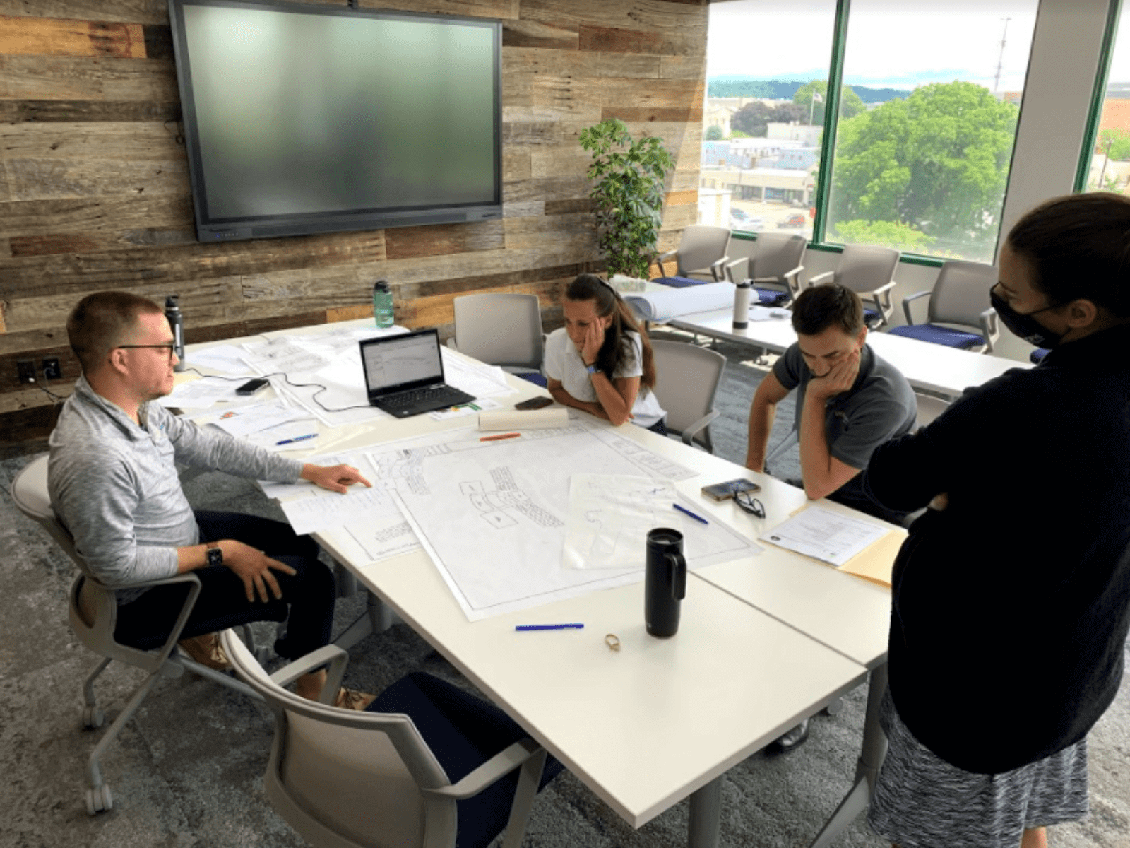 A team of civic architects reviewing a blueprint sitting at a table in a room with a large tv on the wall.
