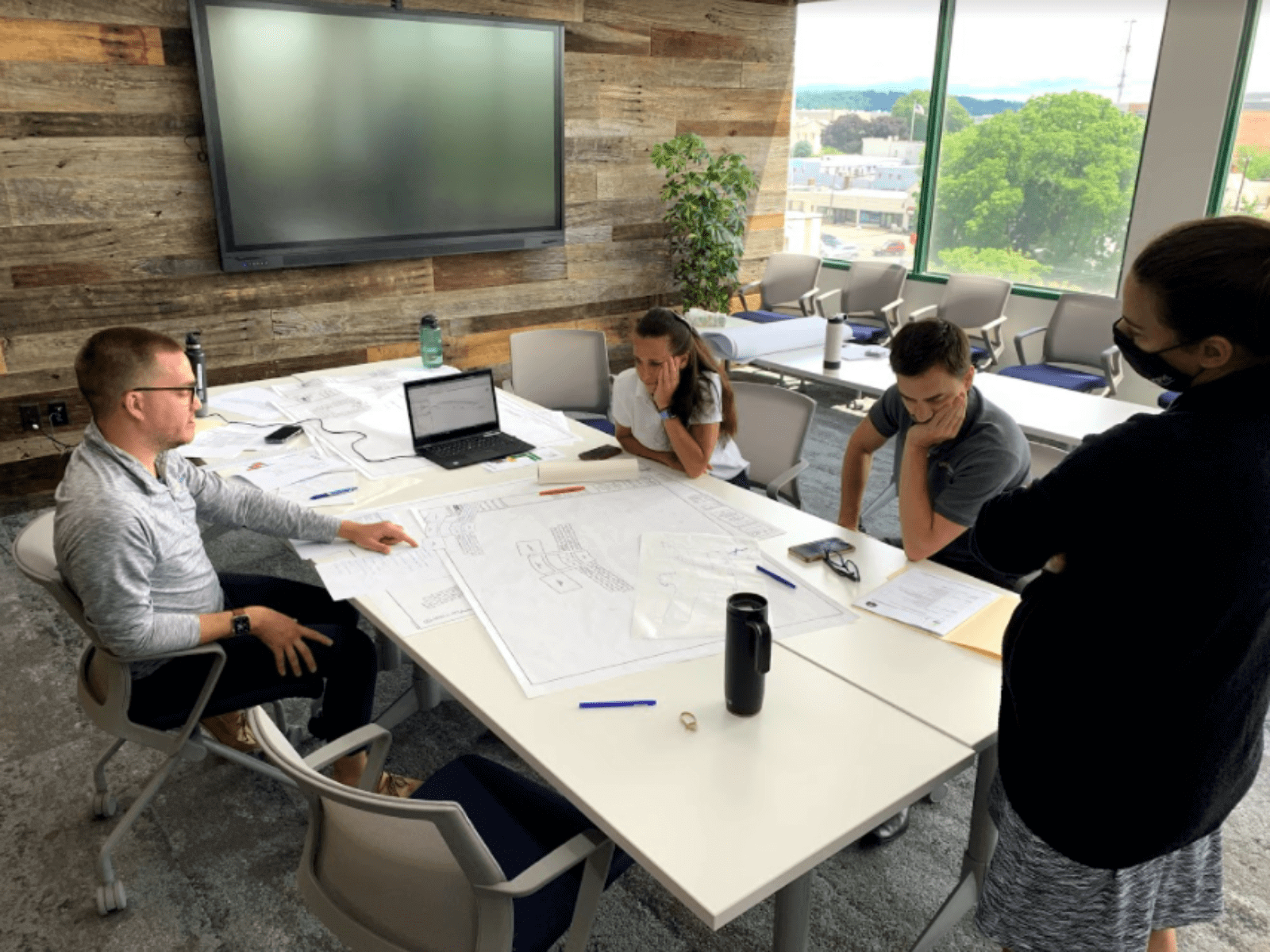 Temp-1600x1200 (1) A team of civic architects reviewing a blueprint sitting at a table in a room with a large tv on the wall.