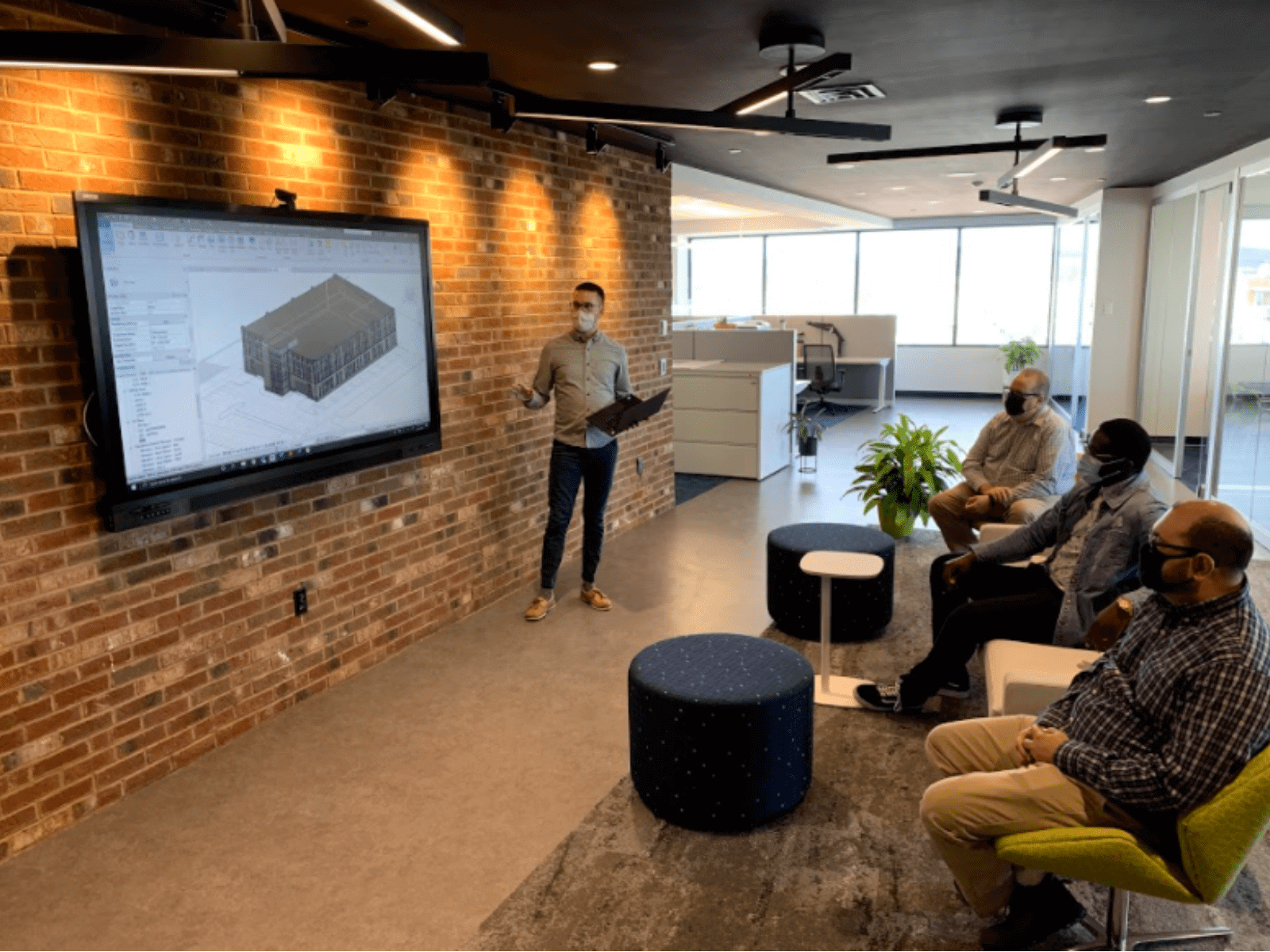 Temp-1600x1200 Three people wearing facemasks listening to another person wearing a face mask give a presentation at an office with a brick wall and plants in the room.
