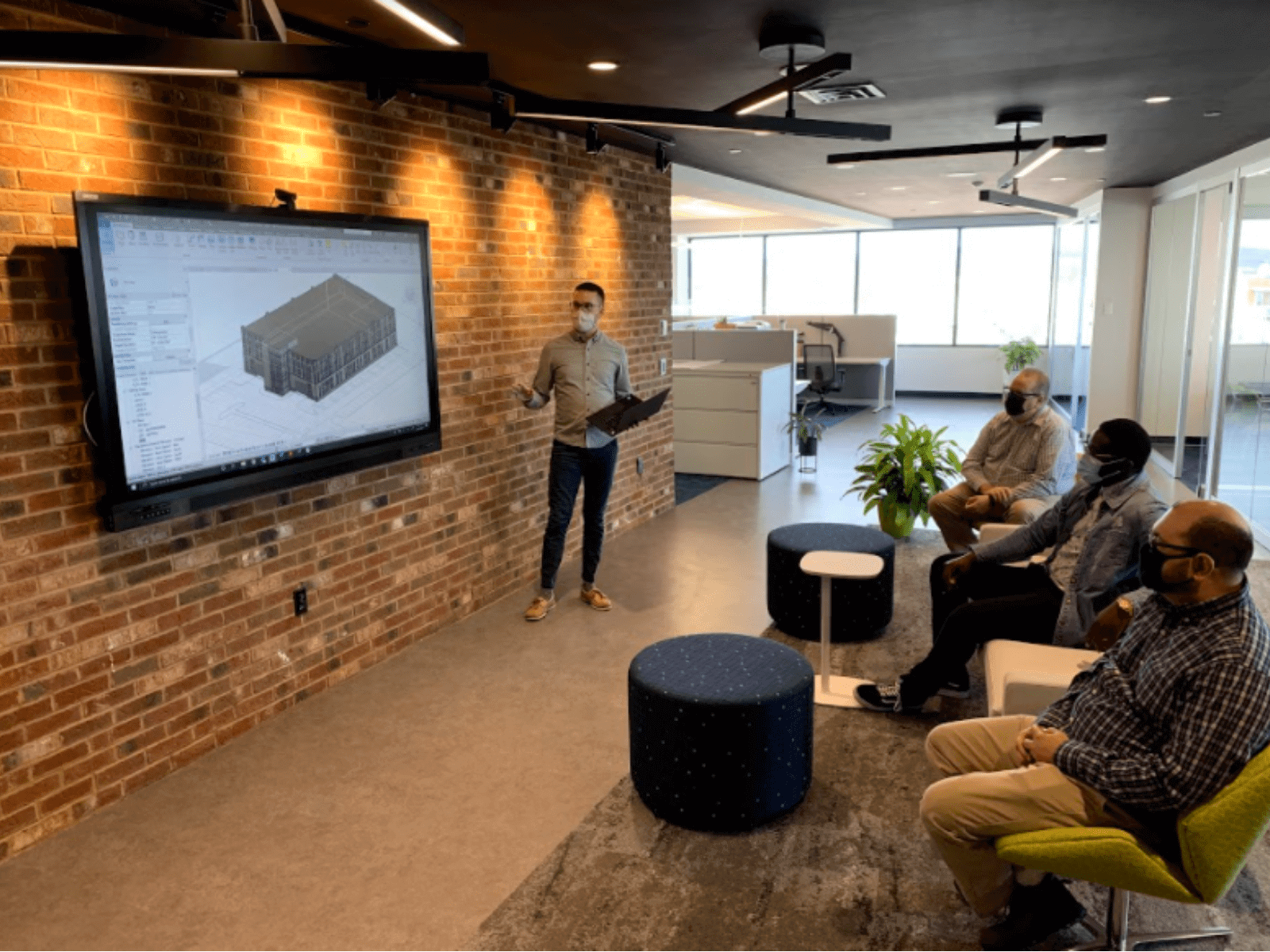 Three people wearing facemasks listening to another person wearing a face mask give a presentation at an office with a brick wall and plants in the room.