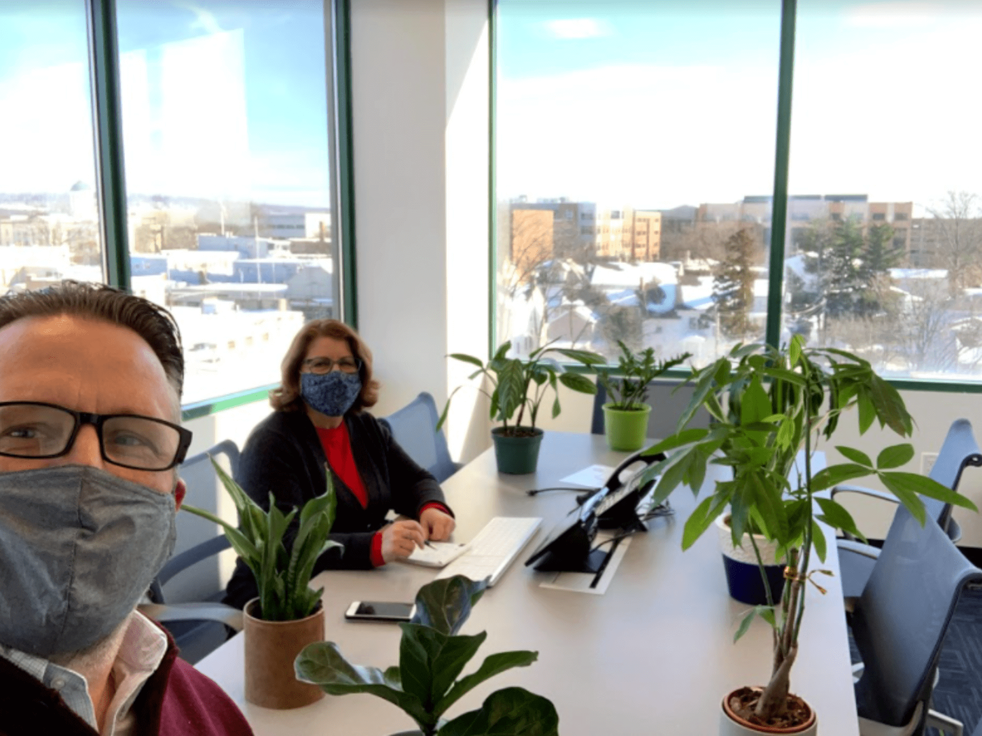 Two people wearing glasses and facemasks in a conference room with big windows and multiple plants on a table