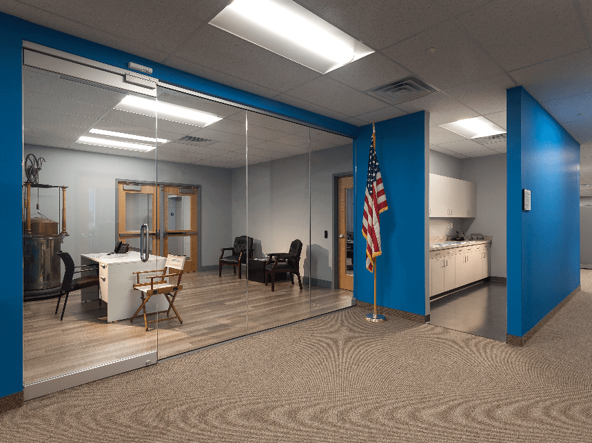An office space with blue walls, an office with clear doors, an american flag, and a kitchen.