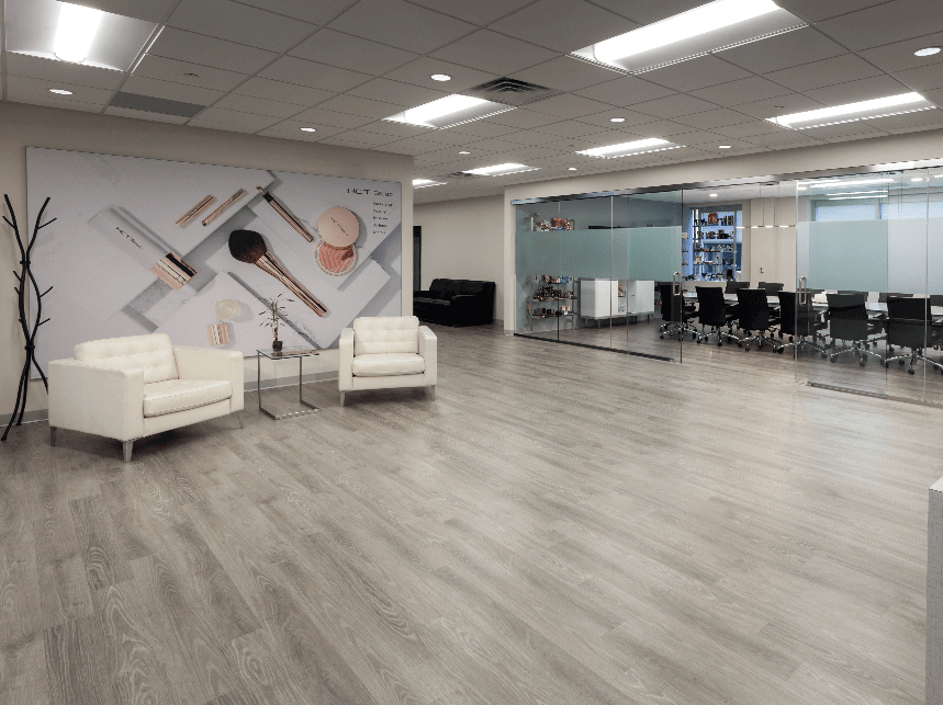 An office space with a large open room with two leather chairs a makeup ad behind the chairs, and a large conference room with many black chairs to the right.