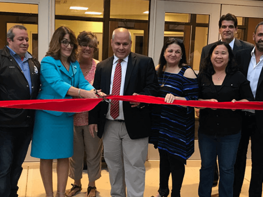 Eight people holding a red ribbon as the woman in the blue cuts the ribbon with a pair of scissors.