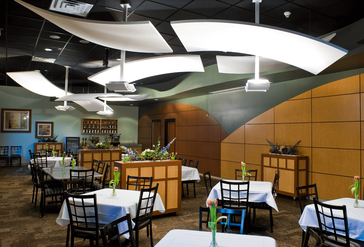 A dining area with many tables that have four chairs, a white table cloth, and a flower in a vase.