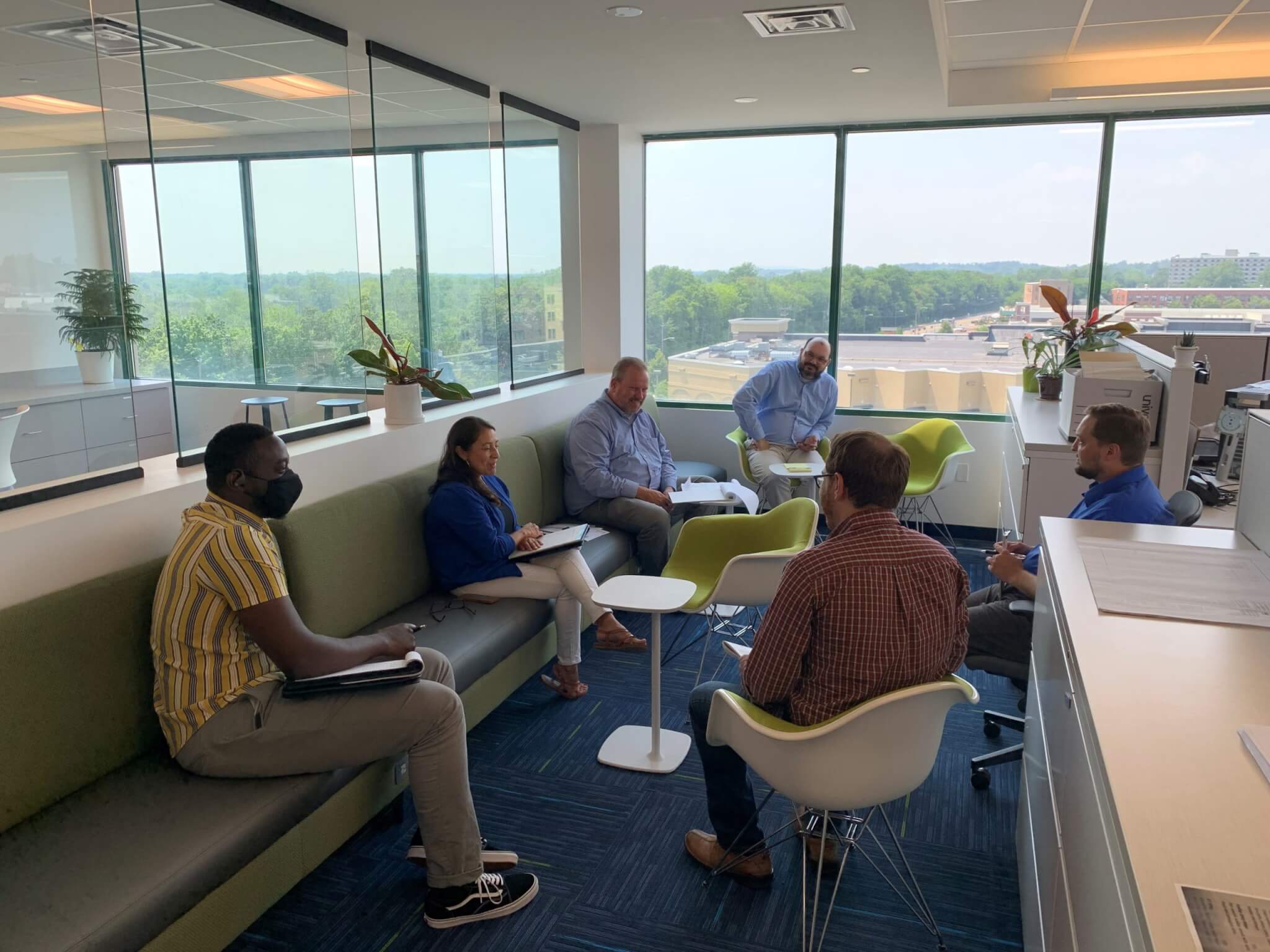 Six people sitting down in an office building having a meeting with plants in the office space.