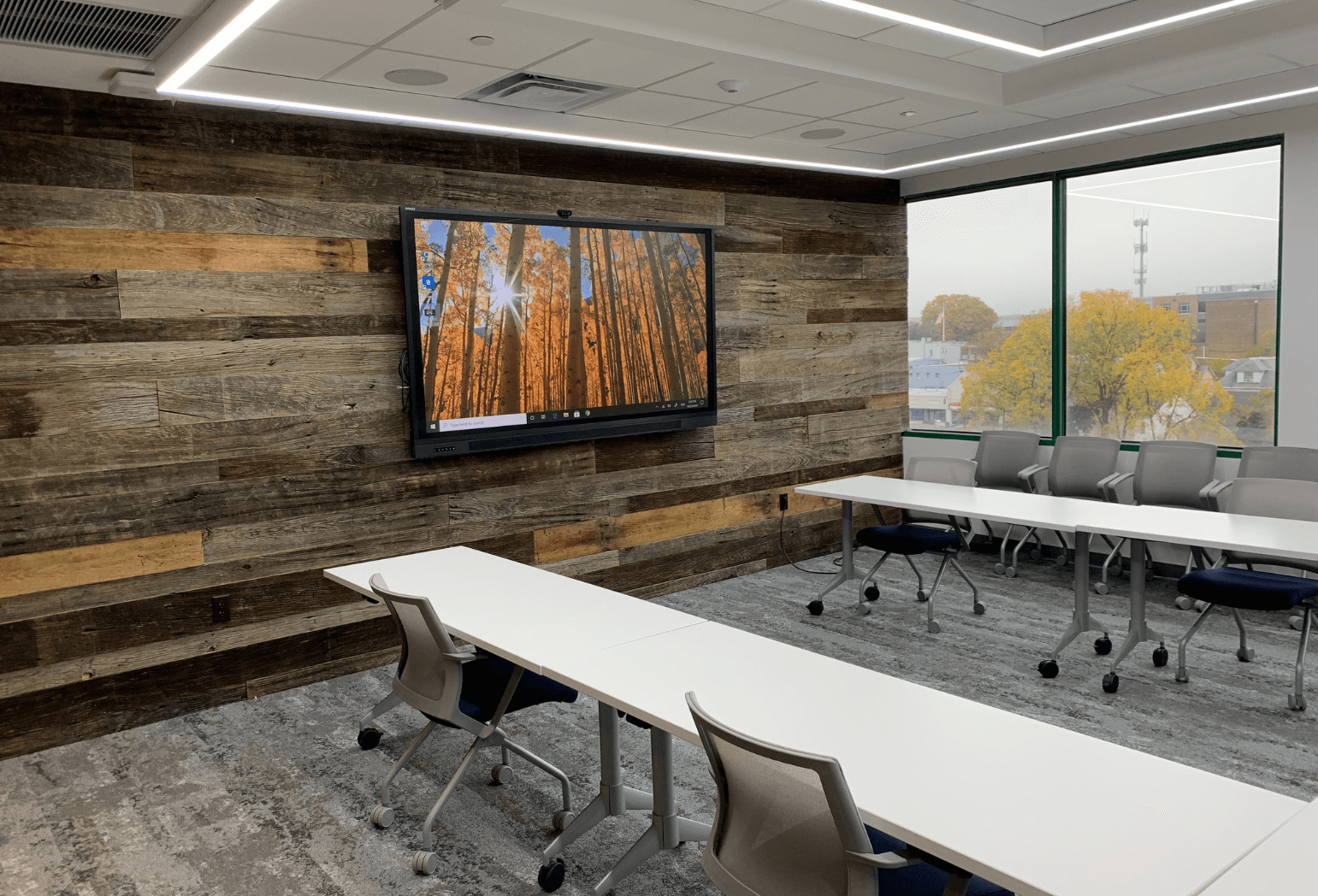A conference room with a wood wall, a tv mounted on the wall, and tables with rolling chairs.